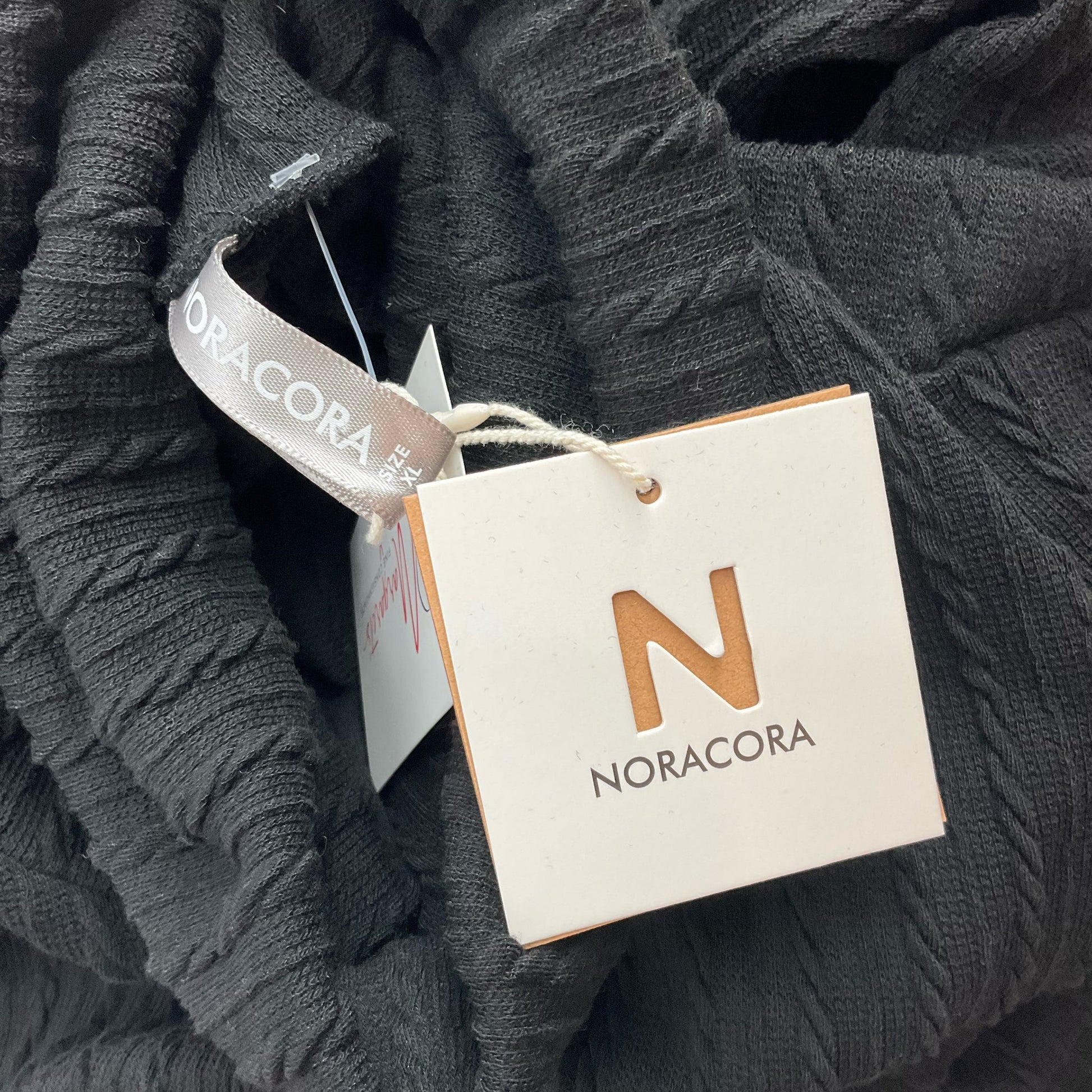 Close-up of Noracora brand tag attached to black ribbed sweater, confirming item is new with tags and consignment-ready.